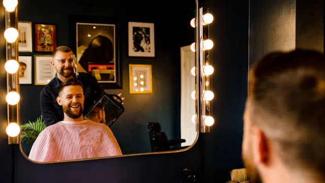 A barber finishing a client’s cut at the bulb-lit mirror